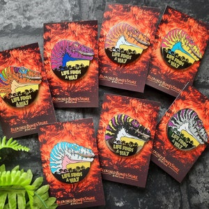 Może przedstawiać: Zestaw dziesięciu szpilek emaliowanych z dinozaurem z napisem "Life Finds a Way" na czerwonym, pomarańczowym i żółtym tle. Szpilki są w różnych kolorach i mają różne wzory.