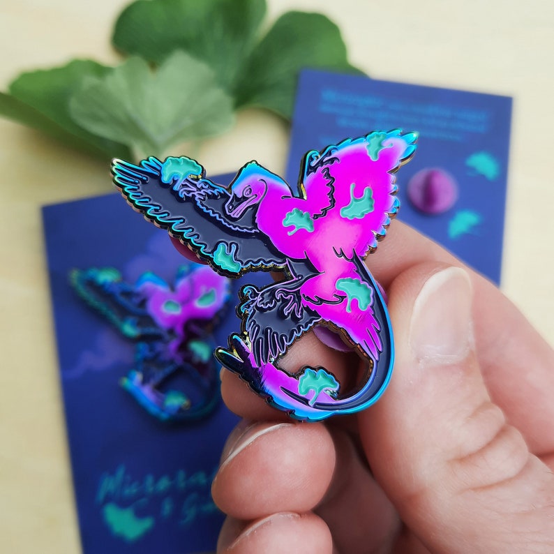 May include: A colorful enamel pin featuring a stylized bird with iridescent wings and a vibrant pink and teal design. The pin is held in a hand, with a matching card in the background. The pin's design includes the text "Aurora & Gaia".