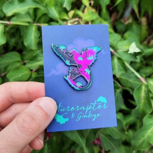 May include: A colorful enamel pin featuring a stylized Microraptor design in shades of pink, black, and teal. The pin is mounted on a blue card with the text "Microraptor & Ginkgo" in teal. The background is a lush green foliage.