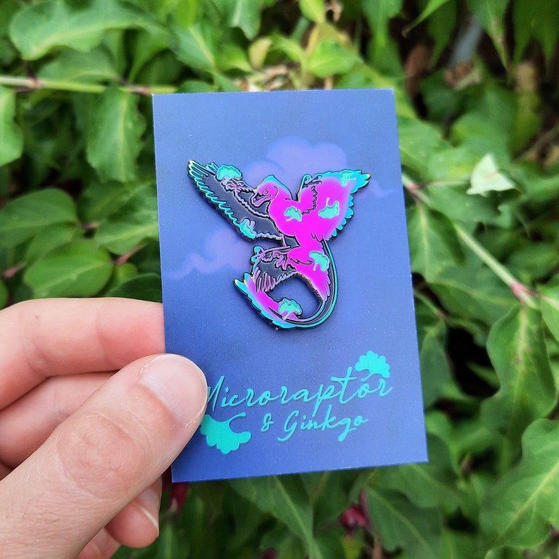 May include: A colorful enamel pin featuring a stylized Microraptor design in shades of pink, black, and teal. The pin is mounted on a blue card with the text "Microraptor & Ginkgo" in teal. The background is a lush green foliage.