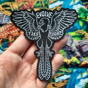 Könnte beinhalten: Schwarzer gestickter Bügeleisen-Aufnäher mit einer weißen Umrandung eines Vogels mit ausgebreiteten Flügeln. Der Text "EVOLVE TAKE FLIGHT" ist um den Vogel herum geschrieben.