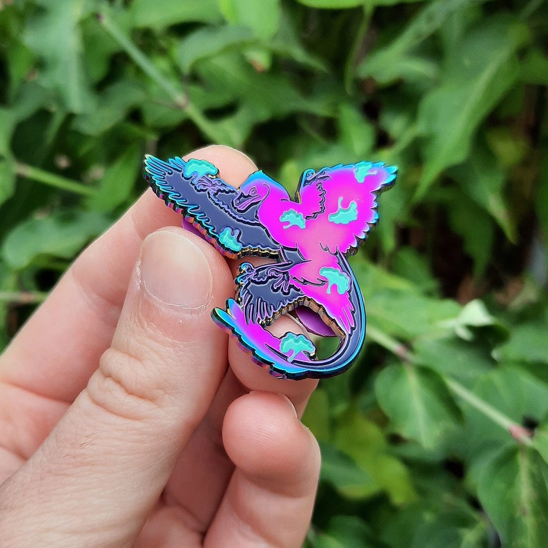 May include: A colorful enamel pin featuring a stylized bird design. The pin has a gradient of pink, purple, and blue hues, with black accents and teal details. The bird's wings are spread, and the pin has a shiny, metallic finish.