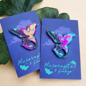 May include: Two iridescent Microraptor pins on blue cards with white cloud designs. The pins feature a colorful design with pink, yellow, and blue hues. The cards have the text "Microraptor & Ginkgo" in a teal color.