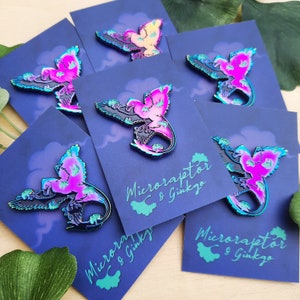 May include: Enamel pins featuring a stylized Microraptor design with a vibrant color scheme of pink, teal, and black. The pins are displayed on dark blue cards with the text "Microraptor & Ginkgo" in a handwritten font.