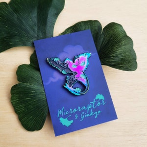 May include: A colorful Microraptor enamel pin with a gradient of pink, blue, and green hues. The pin is set against a dark blue card with the text "Microraptor & Ginkgo" in teal. The design features a stylized dinosaur with outstretched wings.
