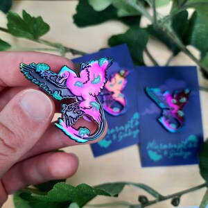 May include: A colorful enamel pin featuring a stylized bird with a vibrant pink and teal gradient. The bird has black wings and is accented with teal and pink details. The pin is held in a hand, with two similar pins on blue cards in the background.
