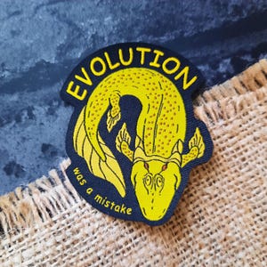 Könnte beinhalten: Gestickter Aufnäher mit einer gelben Cartoon-Kreatur und dem Text "EVOLUTION was a mistake" in Gelb auf marineblauem Hintergrund. Der Aufnäher befindet sich auf einer Jutefläche.
