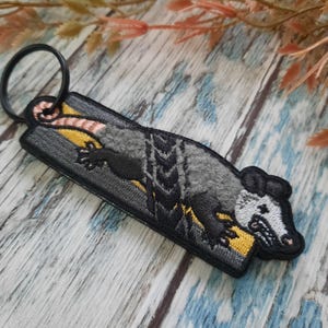 Op de afbeelding: Een grijze en zwarte geborduurde opossum sleutelhanger met een gele streep. De opossum is afgebeeld met een gekrulde staart en een wit gezicht.