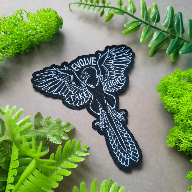 May include: Black embroidered patch with a white outline of a bird with the text "Evolve Take Flight" around it.