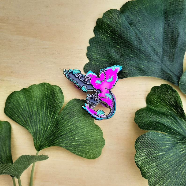 May include: A colorful enamel pin featuring a stylized bird design. The pin showcases a vibrant color palette of pink, teal, and black, with a gradient effect. The bird's wings are spread, and the design has a glossy finish.