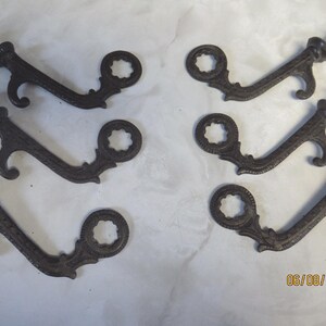 May include: Six antique, dark brown metal shelf brackets. Each bracket has a decorative, openwork circular design and a threaded rod with a nut for mounting. The brackets are arranged on a light-colored surface.