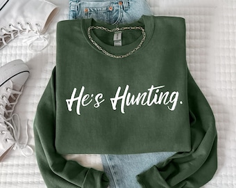 May include: An olive green sweatshirt with the text "He's Hunting" in white script. The sweatshirt is paired with light blue denim jeans, a gold chain necklace, and white canvas sneakers. The items are arranged on a white surface.