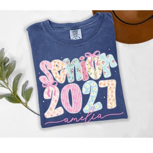 Puede incluir: Una camiseta azul de manga corta con una etiqueta Comfort Colors en el cuello. El frente presenta el texto Senior 2027 en una fuente multicolor con patrones florales y lazos rosas, y el nombre amelia escrito en letra cursiva rosa debajo.