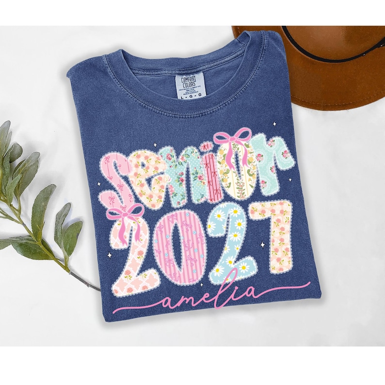 Puede incluir: Una camiseta azul de manga corta con una etiqueta Comfort Colors en el cuello. El frente presenta el texto Senior 2027 en una fuente multicolor con patrones florales y lazos rosas, y el nombre amelia escrito en letra cursiva rosa debajo.