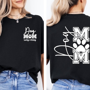 May include: Black t-shirt with white text and graphics. The front features "Dog Mom" with a paw print and the names "Lucky" and "Daisy". The back displays "Love" in cursive, with "MOM" and a paw print graphic.