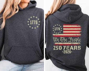 May include: Dark gray hoodie with a front pocket and a hood. The front features the text "1776" surrounded by stars. The back displays a distressed American flag design with the text "We The People" and "250 YEARS -2026".