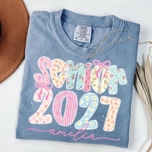 Puede incluir: Una camiseta azul de manga corta con una etiqueta Comfort Colors en el cuello. El frente presenta el texto Senior 2027 en un patr&oacute;n de parches florales y rayas con lazos rosas, y el nombre amelia en letras cursivas rosas bajo los n&uacute;meros.