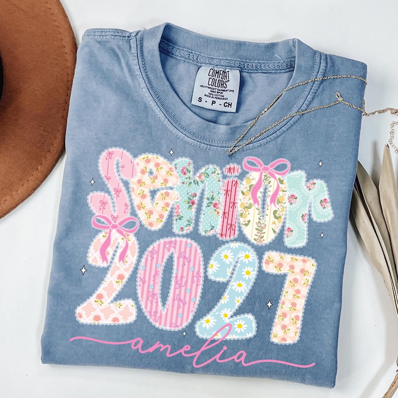 Puede incluir: Una camiseta azul de manga corta con una etiqueta Comfort Colors en el cuello. El frente presenta el texto Senior 2027 en un patr&oacute;n de parches florales y rayas con lazos rosas, y el nombre amelia en letras cursivas rosas bajo los n&uacute;meros.