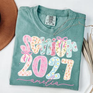 Puede incluir: Una camiseta de manga corta color verde salvia con el texto Senior 2027 en estilo patchwork con patrones florales, de rayas y de cuadros vichy en rosa, azul y amarillo. La palabra amelia est&aacute; impresa en cursiva rosa debajo, con lazos rosas.