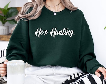 May include: A dark green sweatshirt with the text "He's Hunting" in white script. The sweatshirt is a crew neck style. The image shows a person wearing the sweatshirt, holding a white mug.