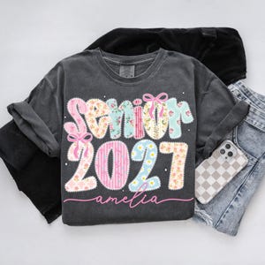 Puede incluir: Una camiseta gris carb&oacute;n de manga corta con un estampado gr&aacute;fico que muestra el texto Senior 2027 en letras grandes de colores pastel con varios patrones, decoradas con lazos rosas y flores, con el nombre amelia en letra cursiva rosa.