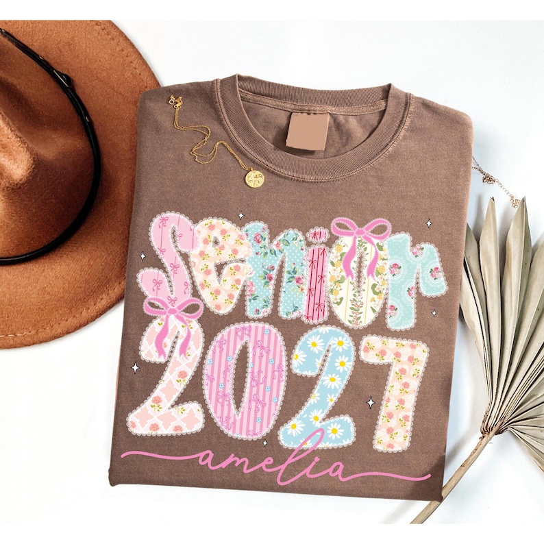 Puede incluir: Una camiseta marr&oacute;n de cuello redondo con el texto "Senior 2027" en letras grandes rellenas de varios patrones florales y de lazos en tonos rosa, azul y amarillo. El nombre "amelia" est&aacute; impreso en cursiva rosa debajo del texto.