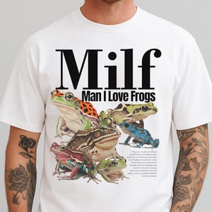 Può includere: T-shirt bianca con una grafica di diverse rane colorate. La maglietta ha la scritta "Milf" in grandi lettere nere sopra "Man I Love Frogs". Le rane sono di vari colori, tra cui verde, arancione e blu.