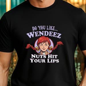 May include: Black t-shirt featuring a cartoon image of a girl with red pigtails and the text "DO YOU LIKE... WENDEEZ NUTS HIT YOUR LIPS" in white and purple. The girl is winking. The shirt is worn by a person.