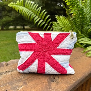 May include: A quilted zippered pouch featuring a red, white, and light blue Union Jack flag pattern. The rectangular fabric accessory has a gold-toned metal zipper pull at the top and visible stitching throughout the quilted surface texture.