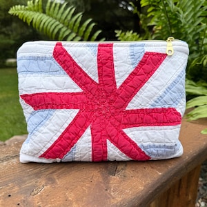 May include: A rectangular quilted zipper pouch featuring a stylized Union Jack design in vibrant red, white, and light blue fabric. The pouch has a gold-toned metal zipper pull at the top right corner and a textured, stitched surface throughout.