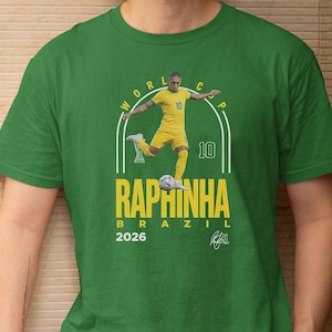 A green t-shirt with a graphic design featuring a soccer player in yellow uniform and the text "WORLD CUP RAPHINHA BRASIL 2026" printed on it.