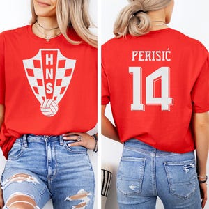 A woman wearing a red t-shirt with a white crest and the number 14 on the back, paired with distressed jeans.