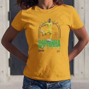 A yellow t-shirt with a graphic design featuring a soccer player in a yellow jersey, along with the text "BAPMINHA BRASIL" and the year "2026".