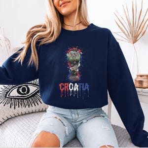 A woman wearing a navy blue sweatshirt with a graphic of a trophy and the word "Crotia" printed on it, sitting on a couch.
