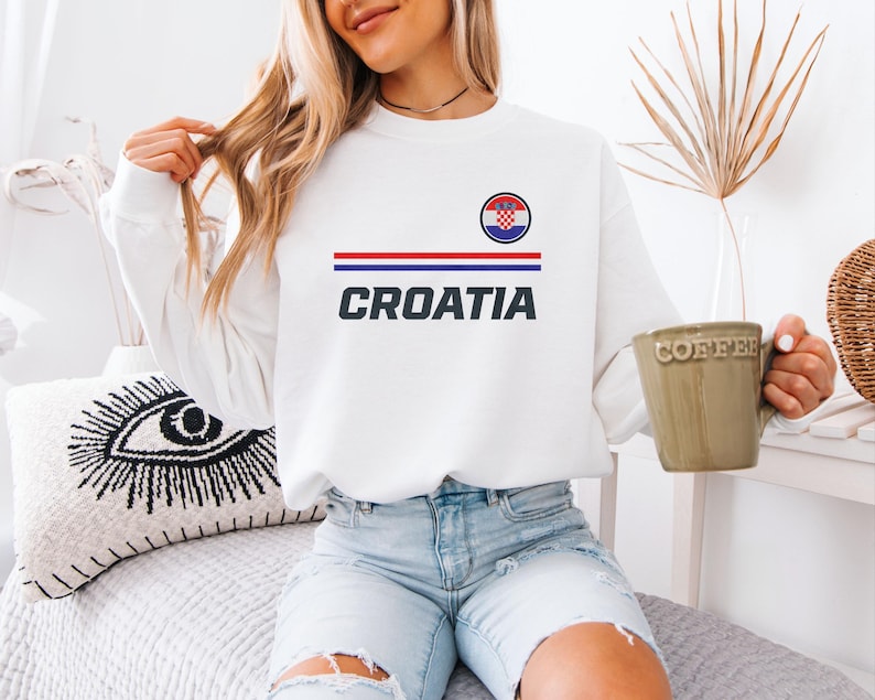 A woman wearing a white sweater with the word "Croatia" printed on it, sitting on a bed and holding a coffee mug.