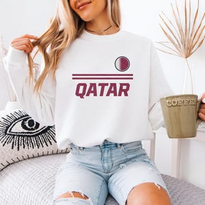 A woman wearing a white sweatshirt with the word "Qatar" printed on it, sitting on a bed and holding a coffee mug.