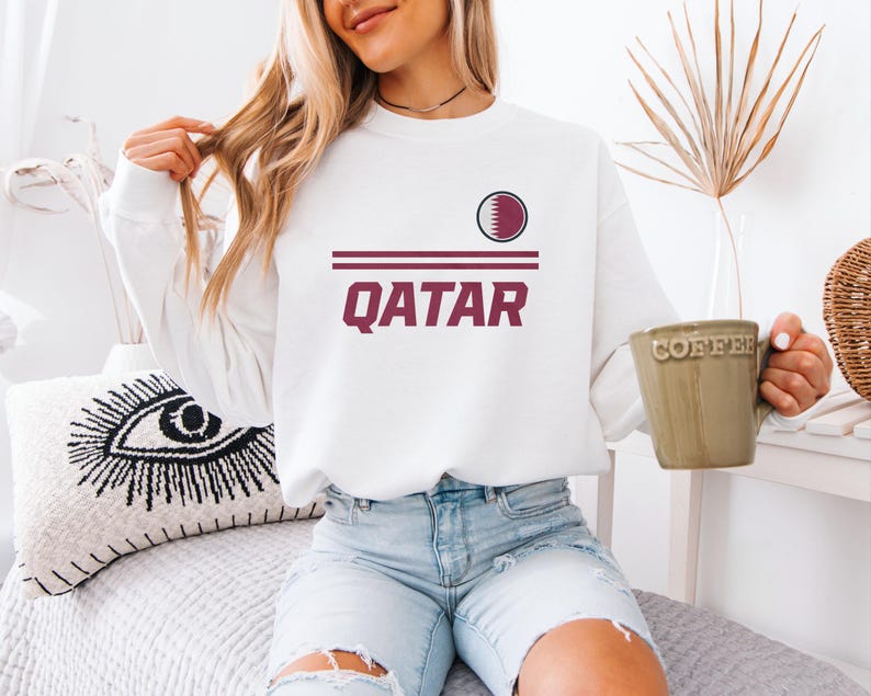 A woman wearing a white sweatshirt with the word "Qatar" printed on it, sitting on a bed and holding a coffee mug.
