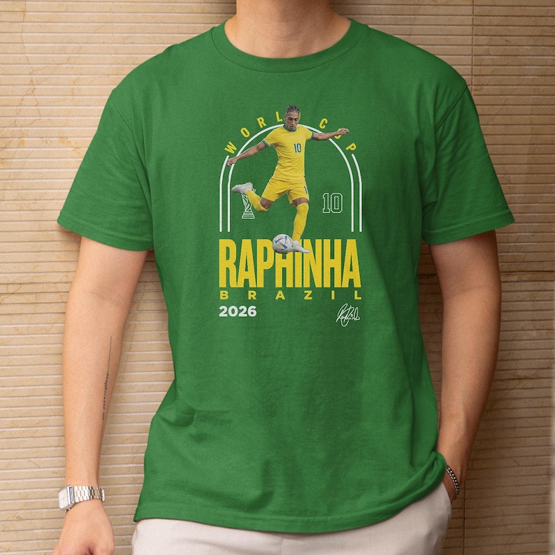 A green t-shirt with a graphic design featuring a soccer player in yellow uniform and the text "WORLD CUP RAPHINHA BRASIL 2026" printed on it.