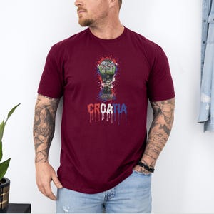 A man wearing a burgundy t-shirt with a graphic of a trophy and the word "CRAZIA" printed on it, standing in front of a white wall.