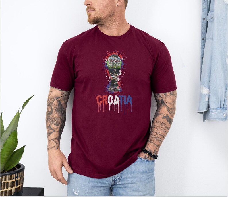 A man wearing a burgundy t-shirt with a graphic of a trophy and the word "CRAZIA" printed on it, standing in front of a white wall.