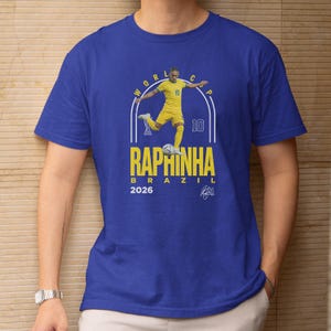 A blue t-shirt with a graphic design featuring a soccer player in yellow uniform and the text "RAPHINHA BRASIL" along with the number "10" and the year "2026".