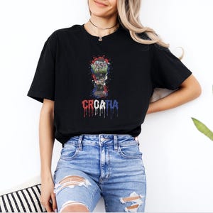 A woman wearing a black t-shirt with a graphic of a trophy and the word "Croatia" printed on it, standing in front of a plant.