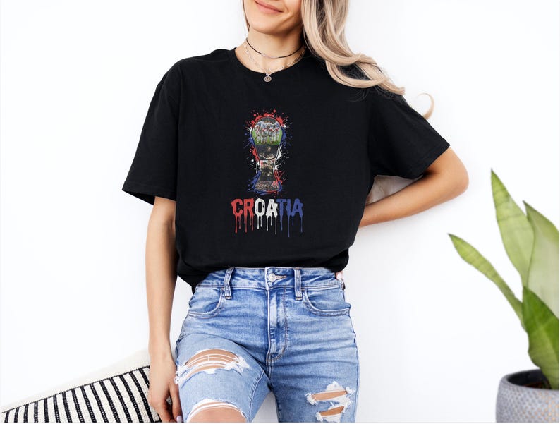 A woman wearing a black t-shirt with a graphic of a trophy and the word "Croatia" printed on it, standing in front of a plant.
