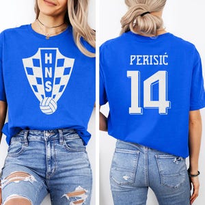 A woman wearing a blue t-shirt with a white logo and the number 14 on the back, paired with distressed jeans.