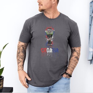 A man wearing a gray t-shirt with a graphic design of a trophy and the word "Croatia" printed on it, standing next to a plant.