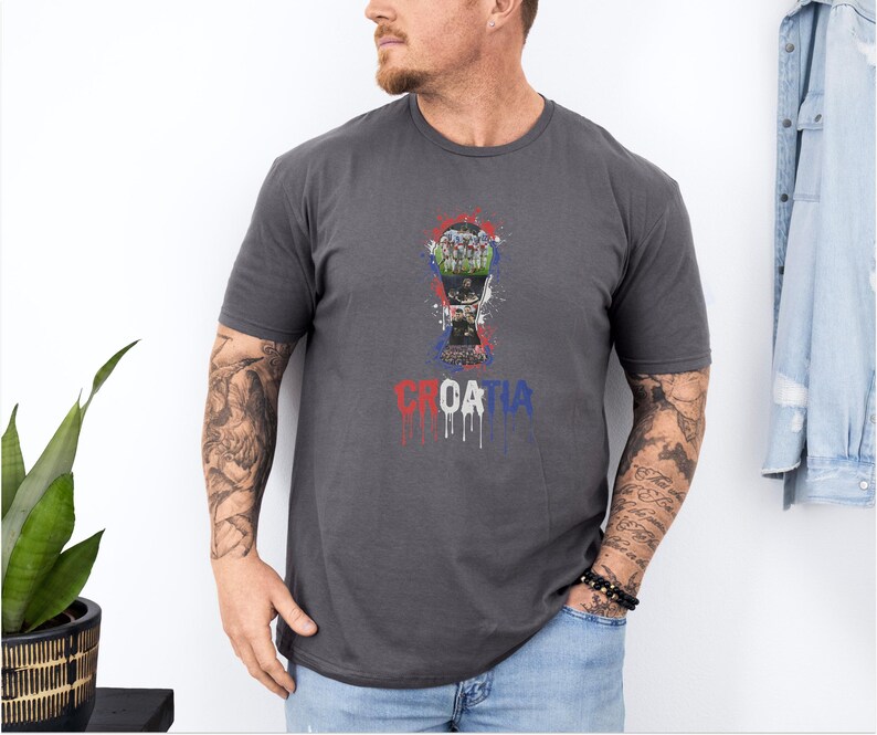 A man wearing a gray t-shirt with a graphic design of a trophy and the word "Croatia" printed on it, standing next to a plant.