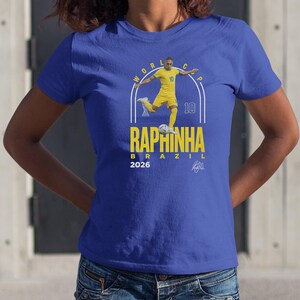 A blue t-shirt with a graphic design featuring a soccer player in yellow and white, along with the text "RAPINHA" and "2026".
