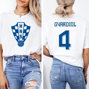 A woman wearing a white t-shirt with a blue shield logo and the number 4 on the back, paired with distressed blue jeans.