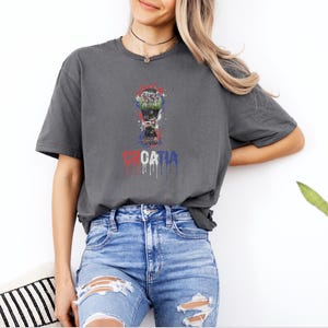 A woman wearing a grey t-shirt with a graphic of a trophy and the word "Croatia" printed on it, standing against a white wall.