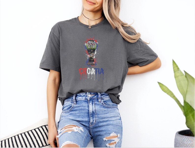 A woman wearing a grey t-shirt with a graphic of a trophy and the word "Croatia" printed on it, standing against a white wall.